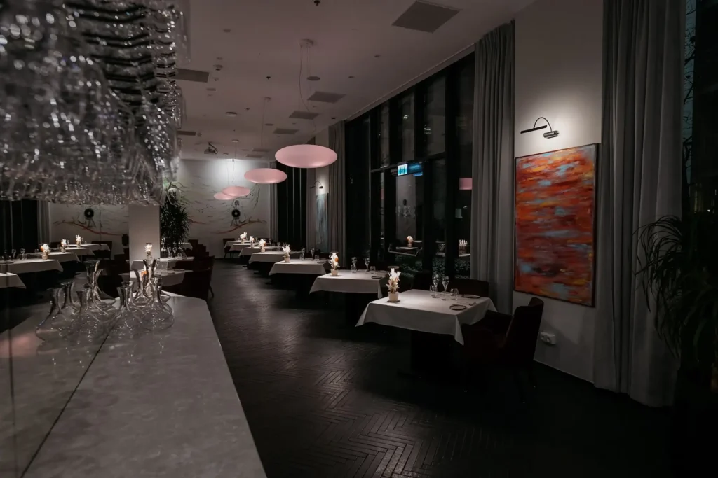 NUTA's Warsaw dining room set for evening service, with white tablecloths, tall windows, pink pendant lights, and hanging glassware above a marble counter.