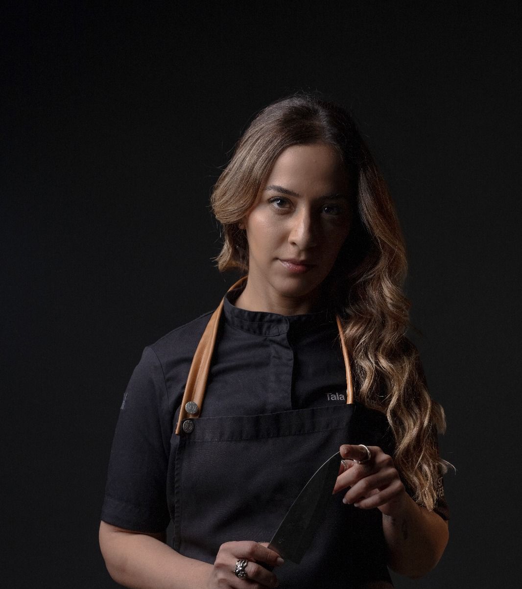 Portrait photograph of Bahraini chef Tala Bashmi in a black chef jacket holding a kitchen knife against a dark background.