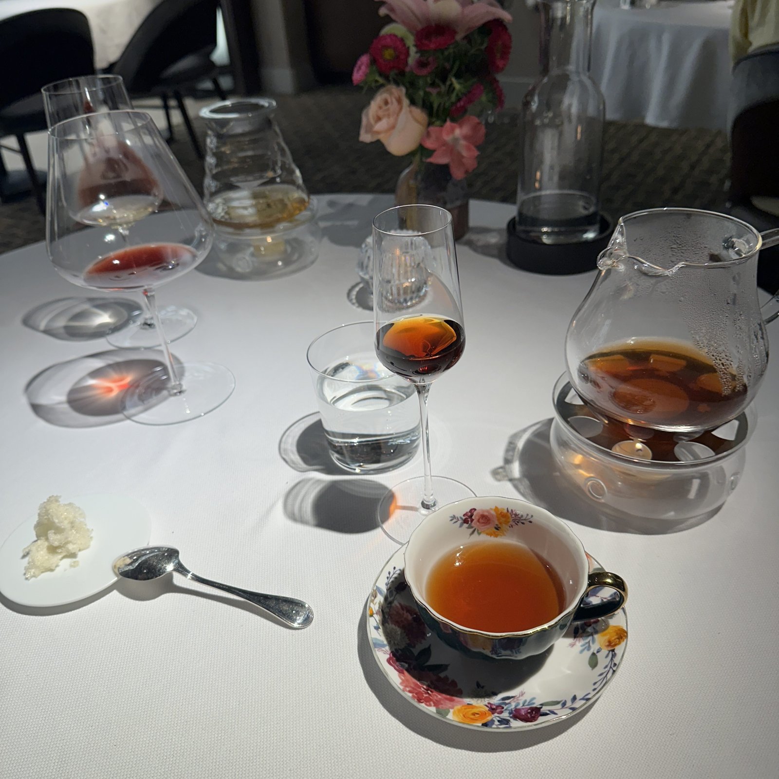 一张 fine-dining 餐桌，花纹茶杯里盛着热茶，高脚杯里有琥珀色茶汤，玻璃茶壶置于保温座上，旁边是水杯与白色桌布服务。
