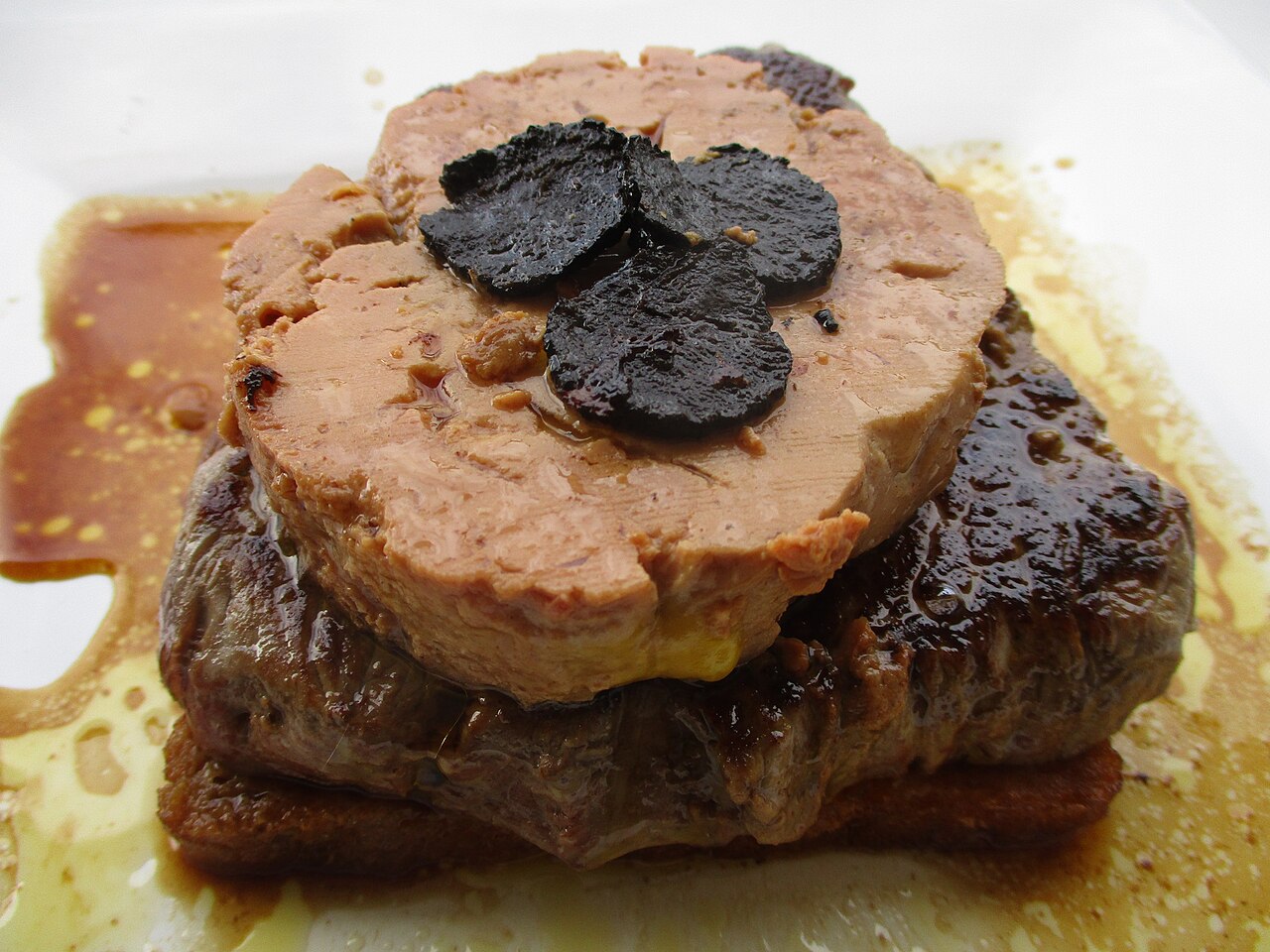 A close photograph of Tournedos Rossini with seared beef, foie gras, truffle slices, sauce, and a browned bread base.