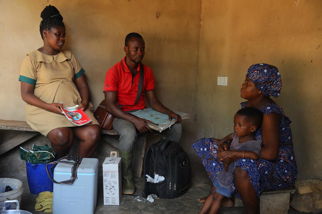 A Ghanaian health worker and volunteer sit with a mother and child during a malaria vaccine community visit, with a cold box and child health materials nearby.