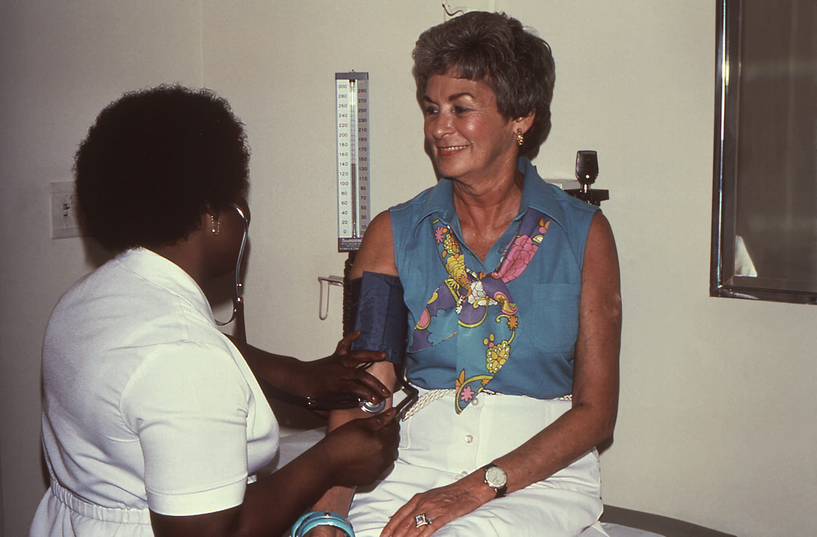 A nurse in 1980 uses a sphygmomanometer and stethoscope to take a seated patient's blood pressure.
