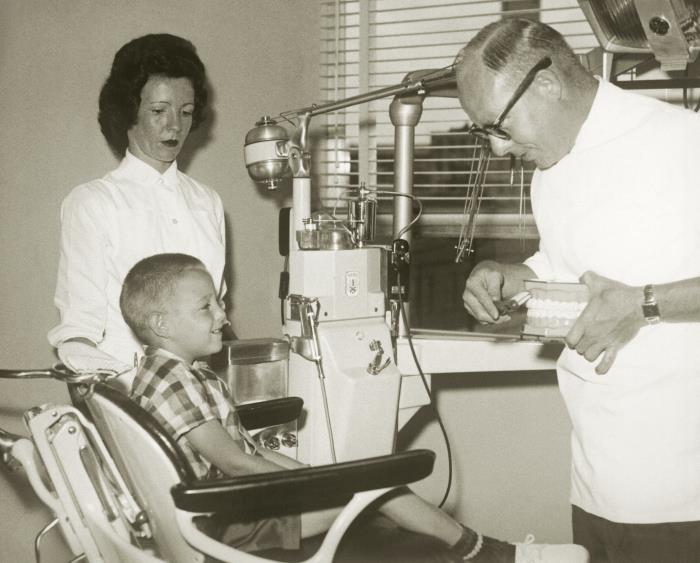 A 1963 CDC archival photograph shows a dentist teaching a young patient how to brush teeth while the child's mother watches.