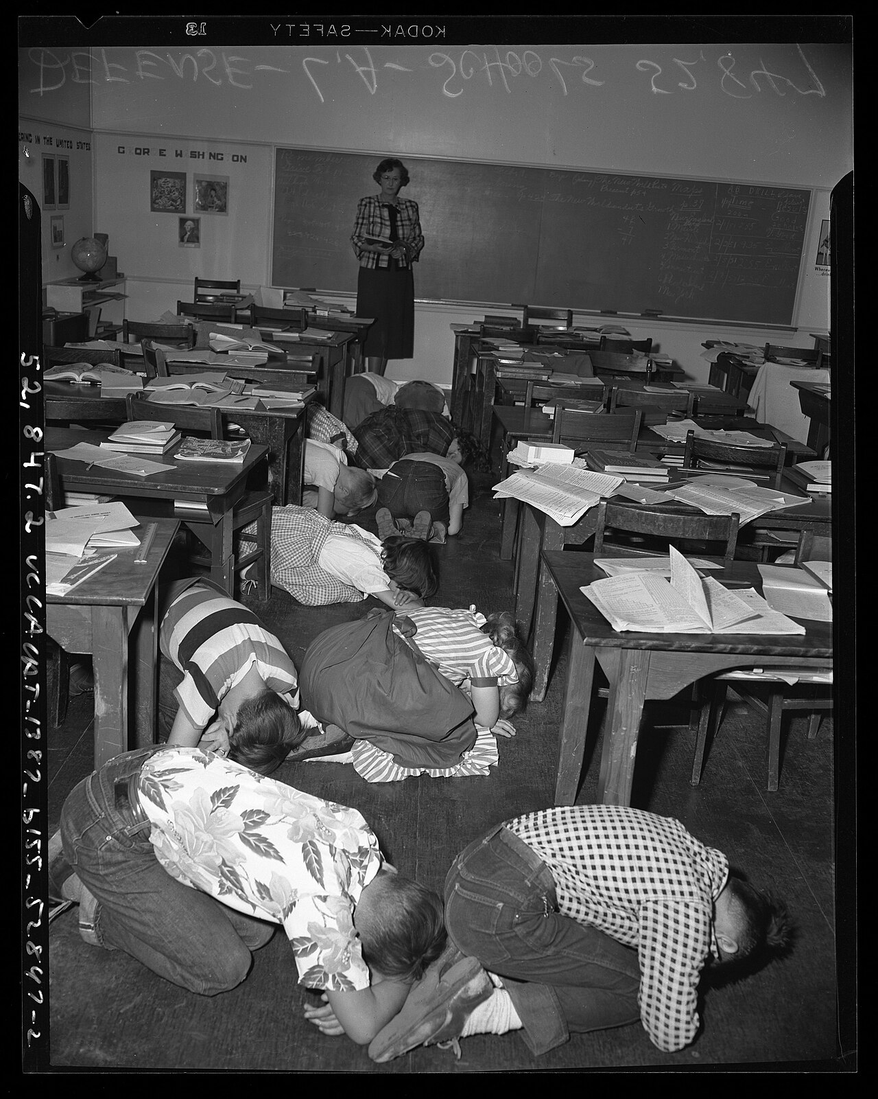 Black-and-white archival photograph of schoolchildren ducking beneath classroom desks during an atom-bomb drill in Los Angeles around 1951.