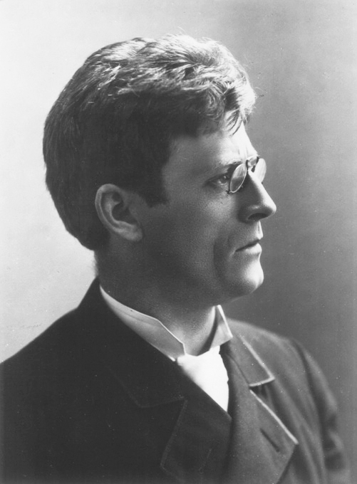 Archival black-and-white photographic portrait of Knut Hamsun around 1890, shown with a mustache and dark suit.