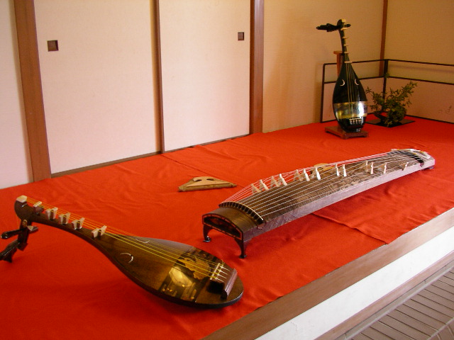 A biwa and koto displayed together in a real room, foregrounding the instrument world behind performed Japanese narrative.