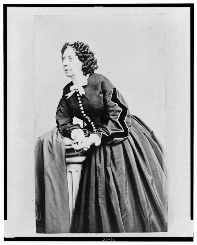 Archival 1866 photographic portrait of Sara Payson Parton, known as Fanny Fern, standing in a dark dress and facing left.