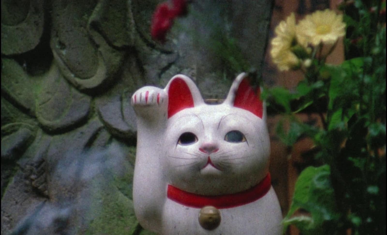 Color film still from Sans Soleil showing a white beckoning cat figurine beside flowers and a carved stone surface.