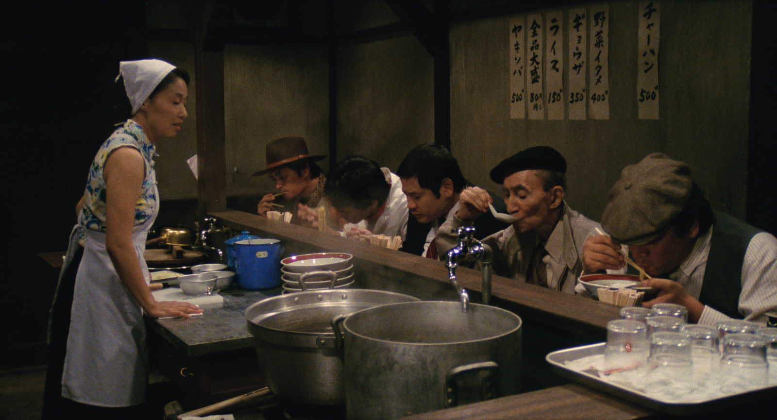 Nobuko Miyamoto as Tampopo stands behind a ramen counter while Goro and the ramen team eat from bowls in Juzo Itami's Tampopo.