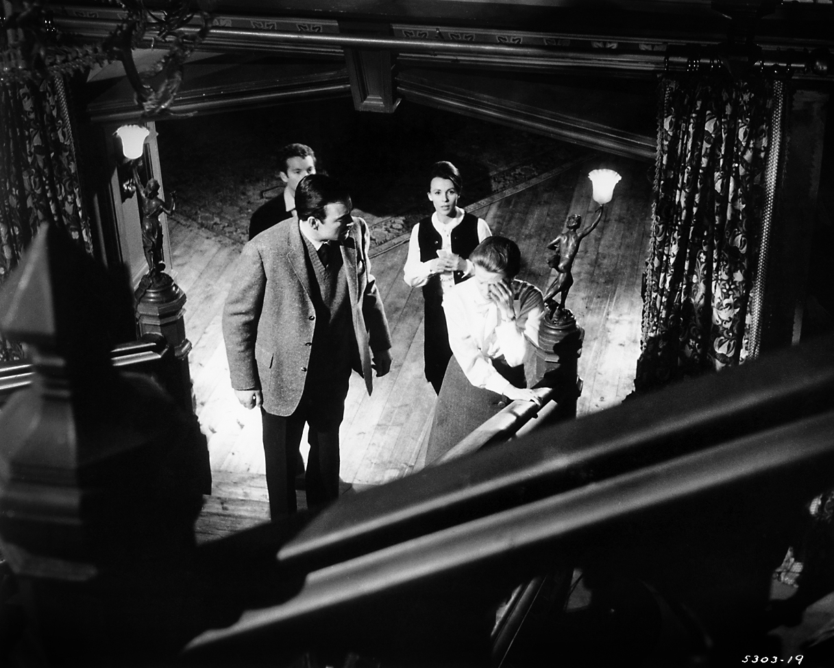 Black-and-white production still from The Haunting showing Eleanor and the investigation group at the foot of Hill House's looming staircase.