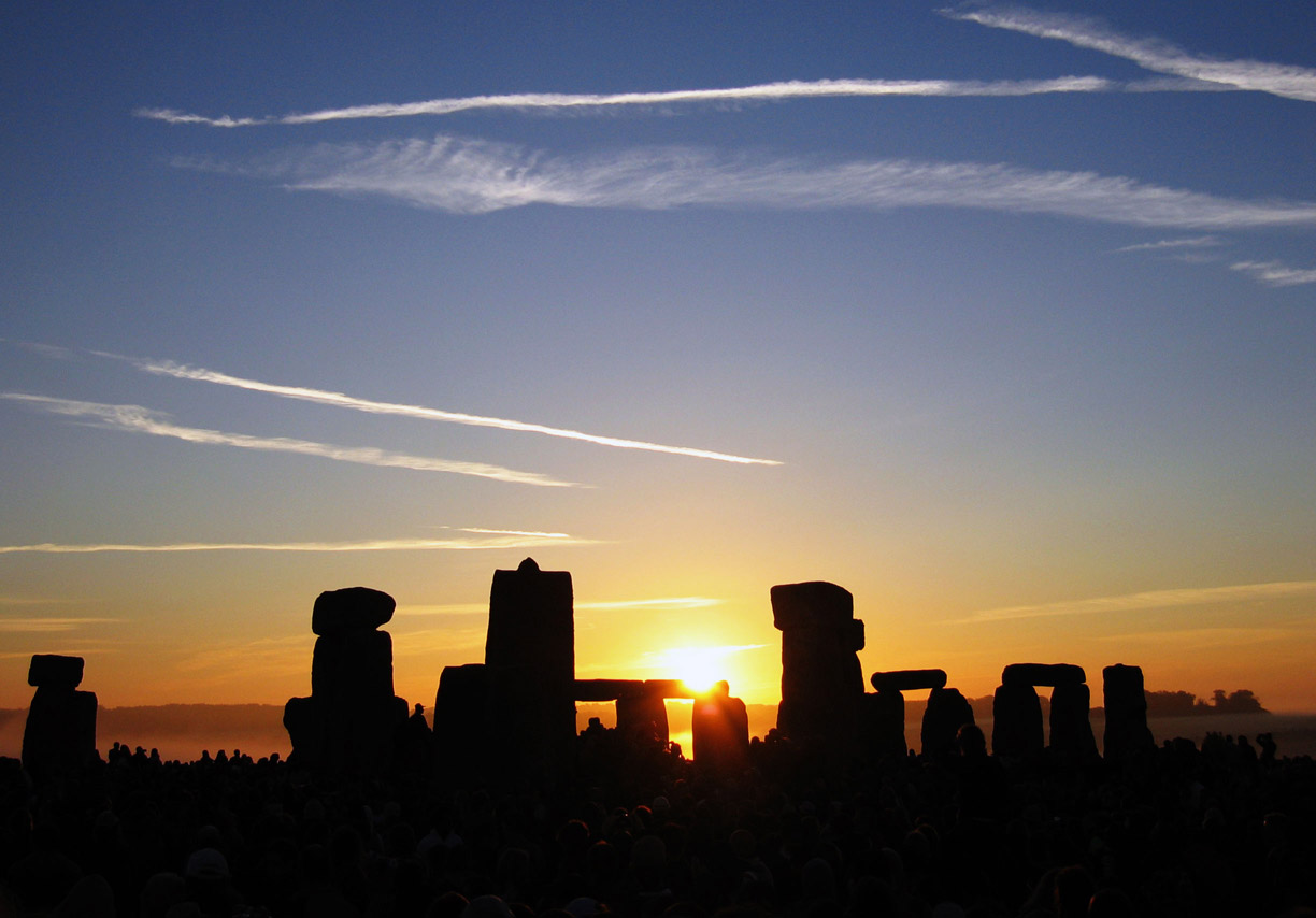 The sun rising behind Stonehenge during the June solstice with a crowd gathered around the stones.