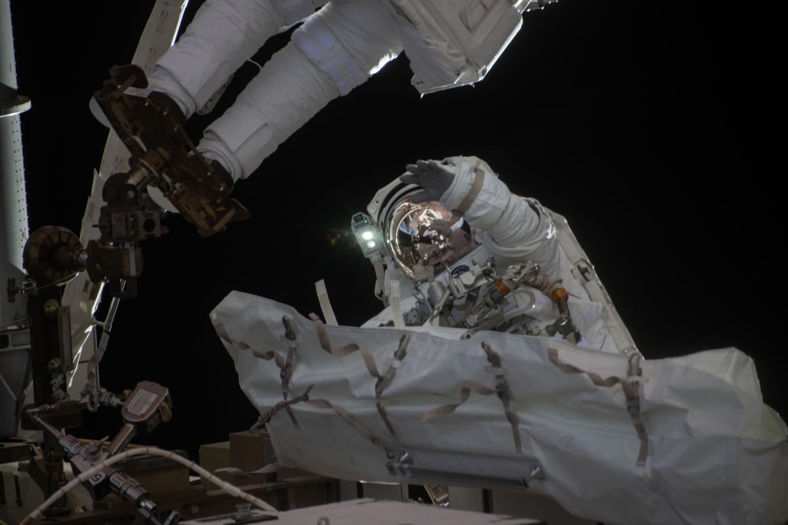 NASA astronaut Jessica Meir waves during a spacewalk outside the International Space Station, with station hardware and Earth visible behind her.
