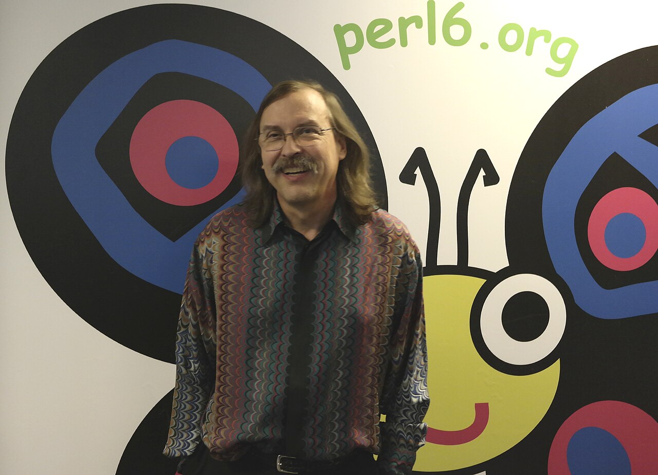 Larry Wall standing at FOSDEM 2015 beside a projected Perl 6 butterfly logo.