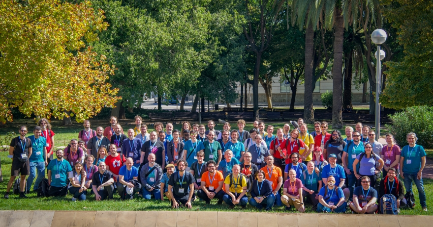 KDE Akademy attendees posing outdoors for a group photograph on grass beneath trees.