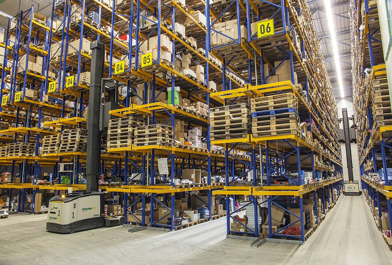 A high-bay warehouse aisle with tall numbered racks, pallets, boxes, and bright overhead lighting.