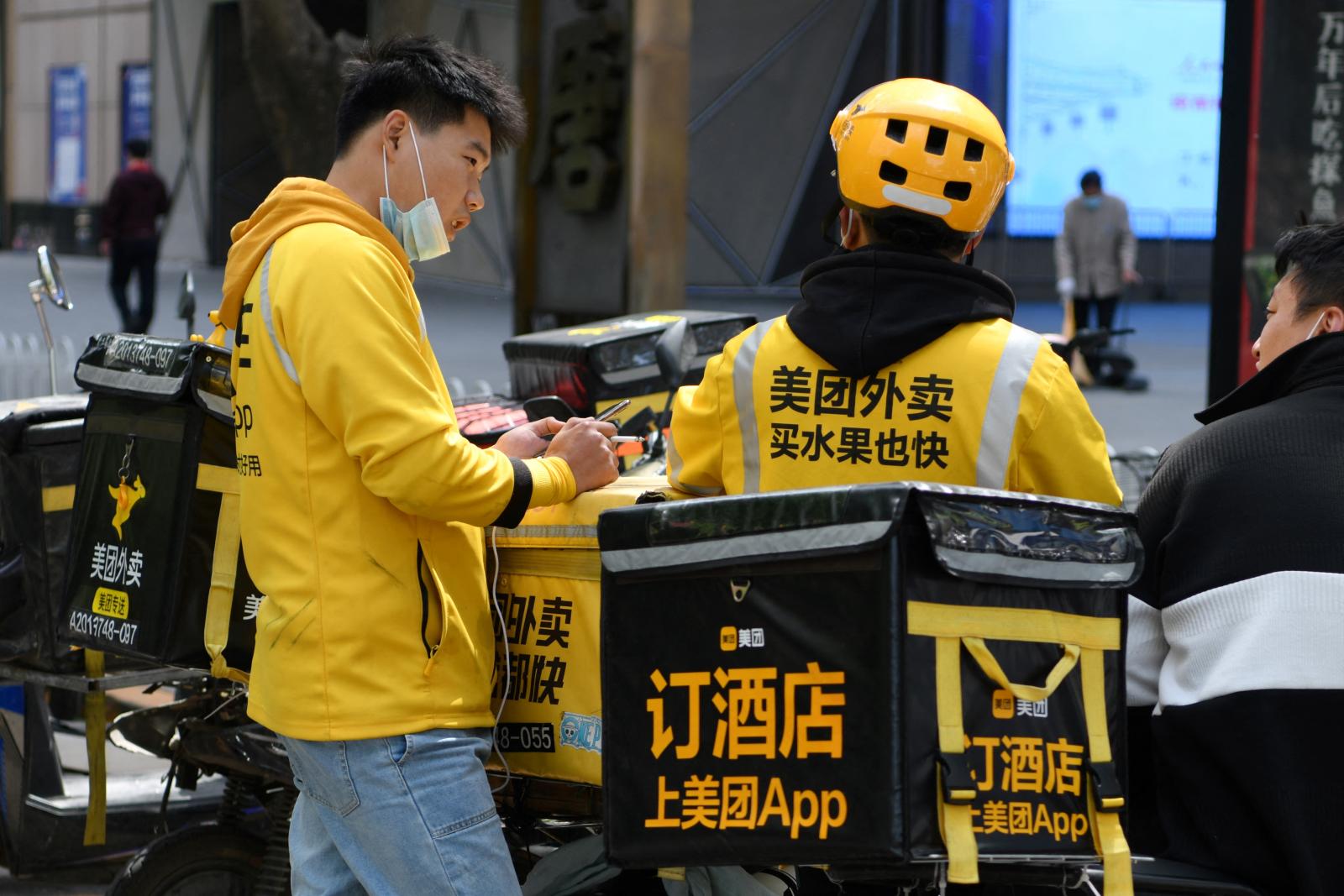 Two Meituan delivery riders in yellow jackets wait beside their motorcycles outside a Beijing restaurant.