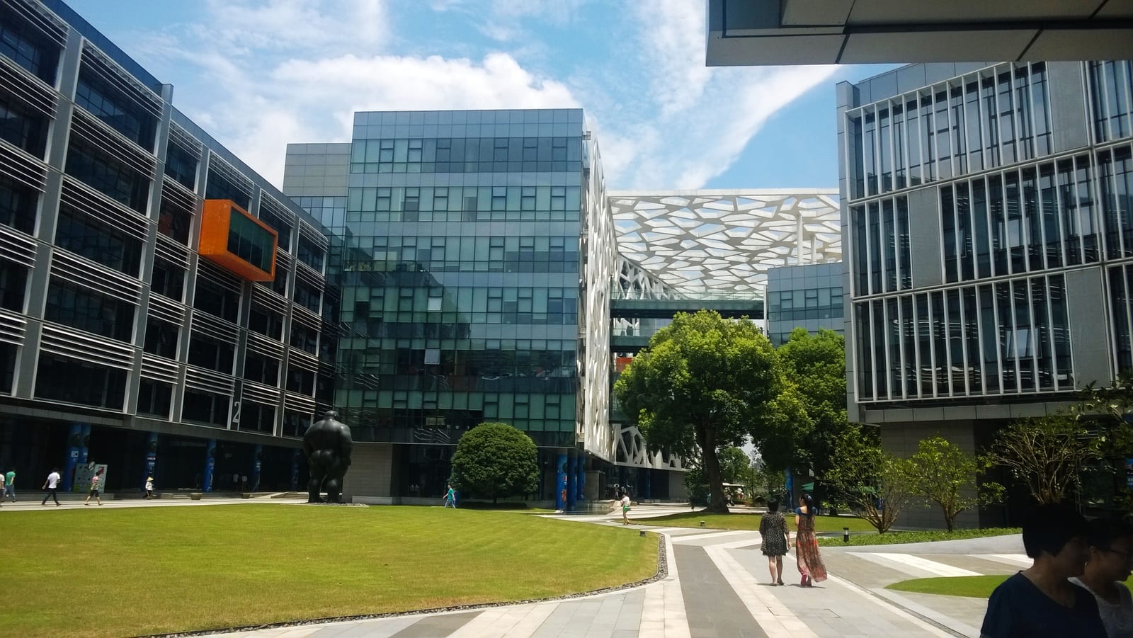 阿里巴巴集团杭州总部园区照片，画面中有玻璃办公楼、草坪、树木，以及穿过庭院的人群。