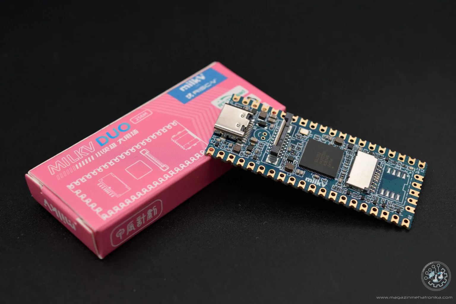 A blue Milk-V Duo 256M development board rests diagonally against its pink retail box on a black tabletop.