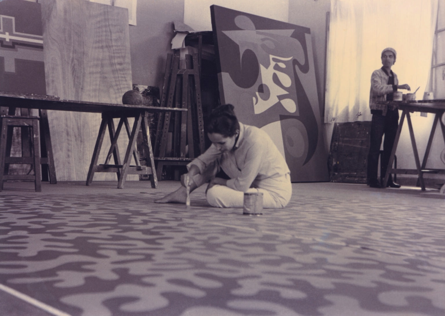 Malika Agueznay sits on a studio floor painting repeated organic forms across a large panel, with geometric paintings and another person behind her.