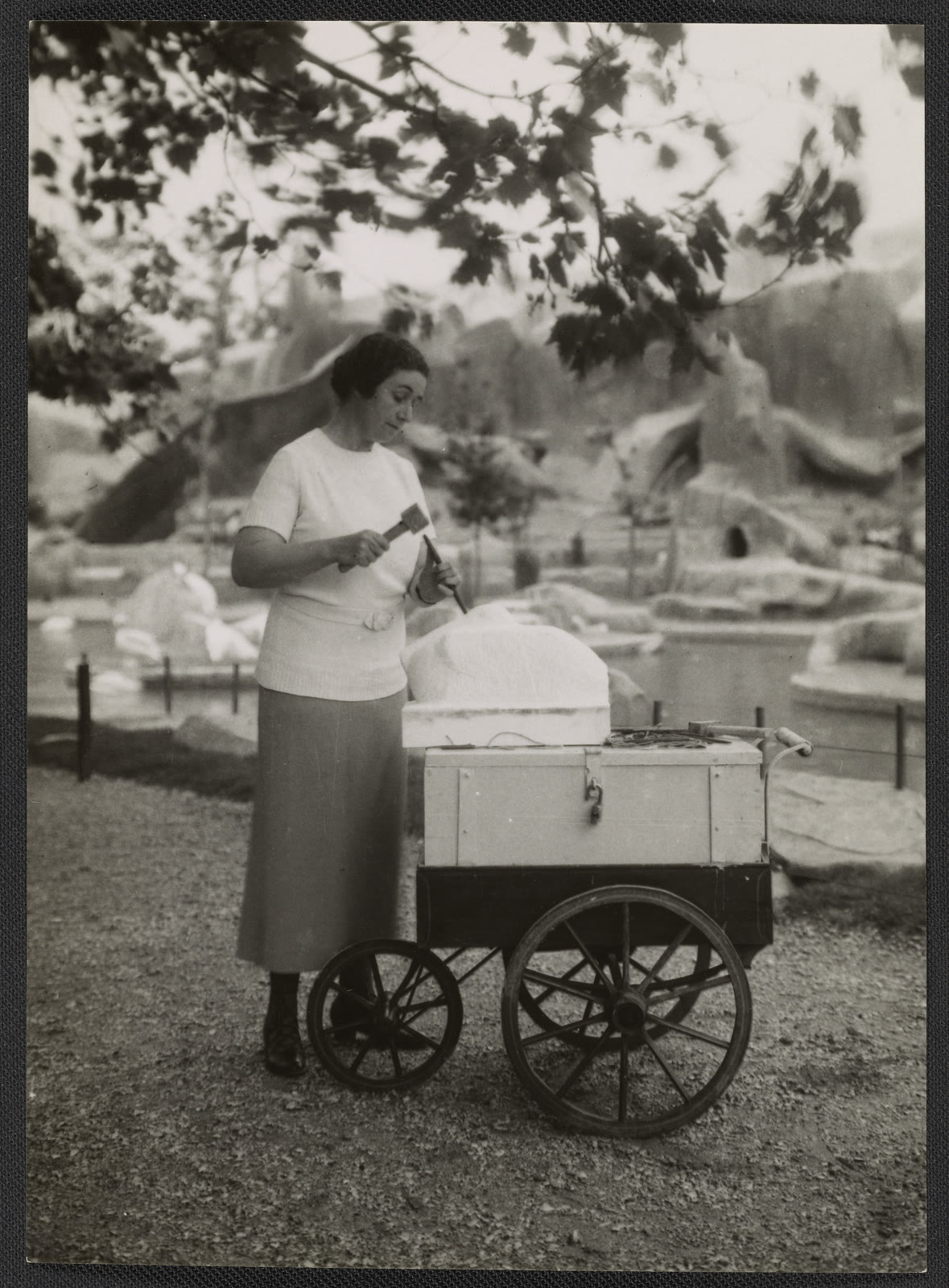 Cornelia Chapin holds a hammer and chisel beside an unfinished pelican sculpture on a large wheeled cart at a Paris zoo.