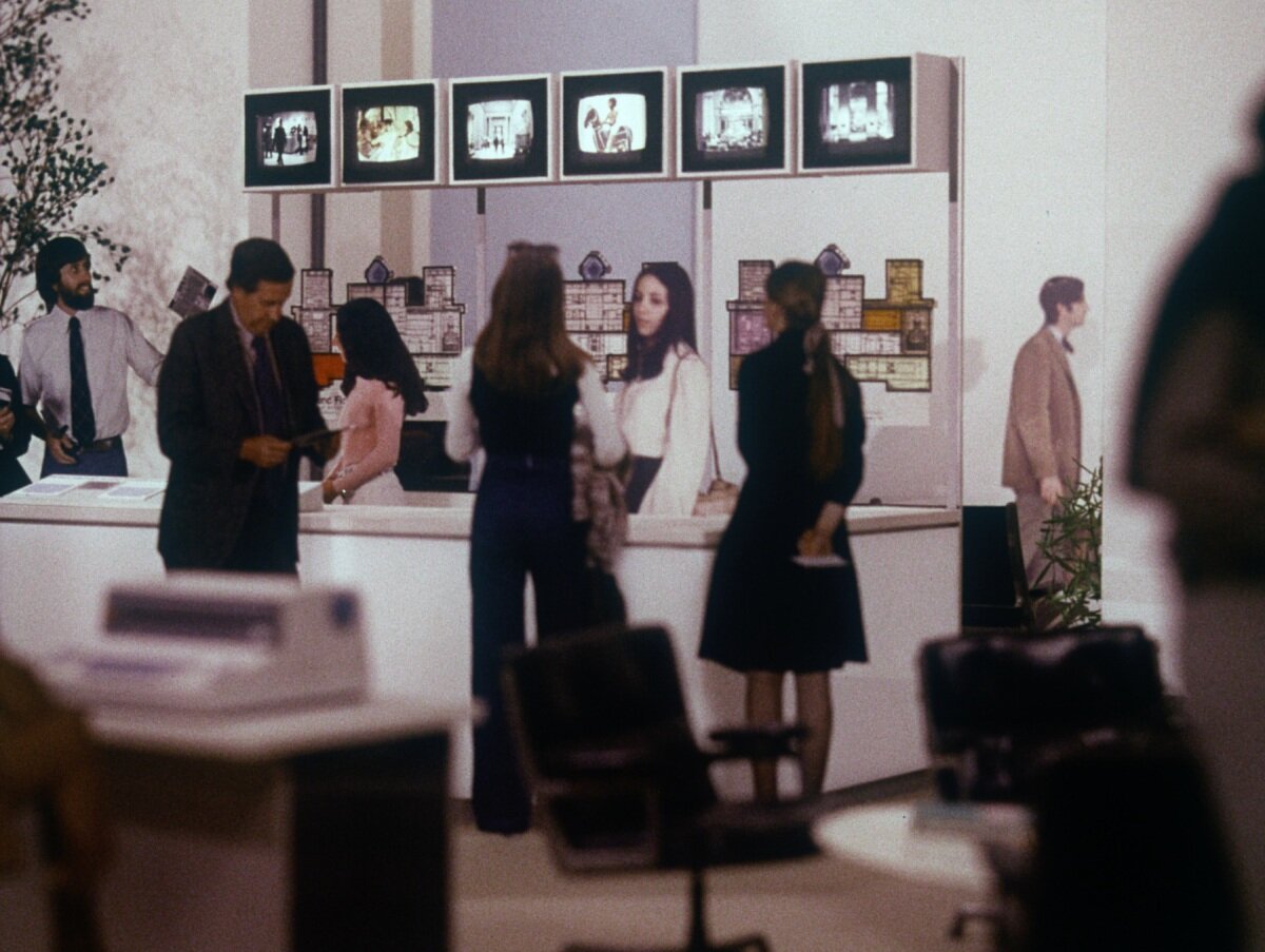 A 1975 color film still shows visitors gathered around an information desk beneath six small video monitors in Charles and Ray Eames's proposed Information Hall for The Metropolitan Museum of Art.