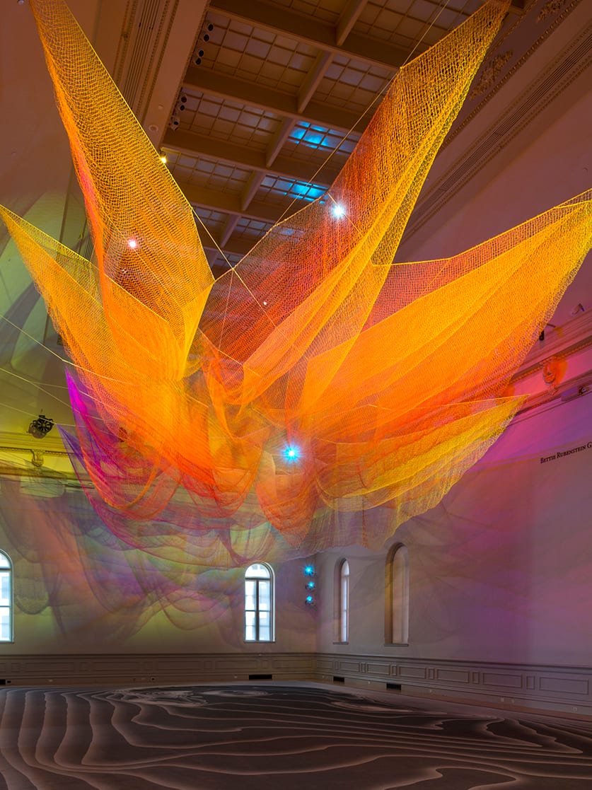 Janet Echelman 一件悬挂的橙黄色网状雕塑，以彩色光影填满 Renwick Gallery 的 Grand Salon。
