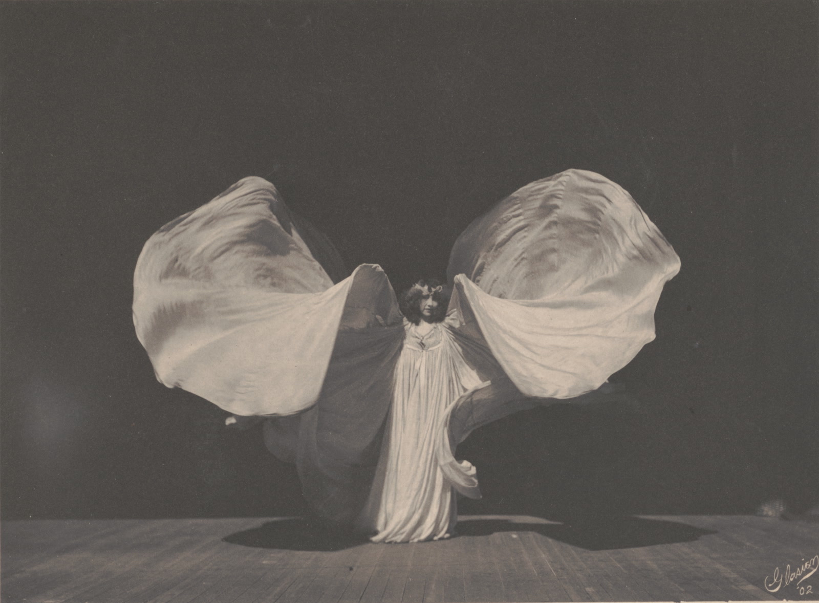 Loïe Fuller faces the camera with both arms raised, spreading a vast pale silk costume into a butterfly-like shape.