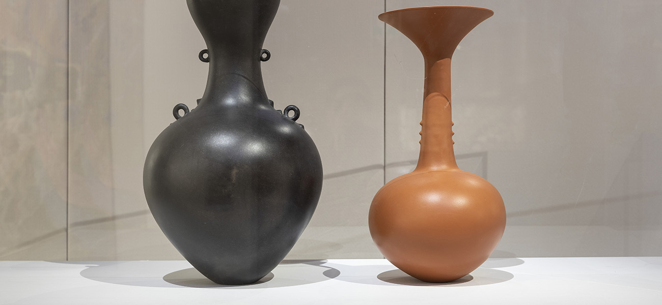 Two Magdalene Odundo ceramic vessels displayed together: a wide black vessel with a rounded body and looped details beside a red-orange vessel with a long neck and flared mouth.
