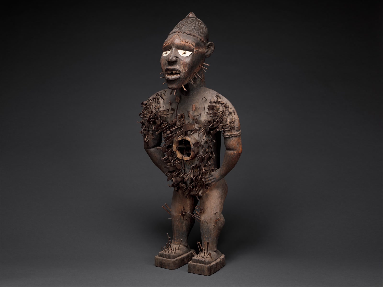 Studio photograph of the Met's nearly four-foot-tall Mangaaka power figure, a dark wooden figure with bright white eyes, hands on hips, dense iron additions across its torso, and an open circular cavity in its abdomen.