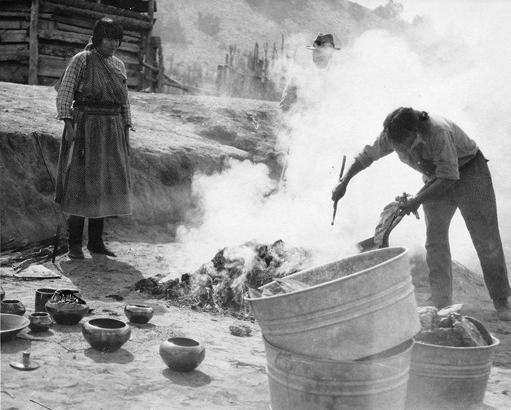 Archival black-and-white photograph of Maria and Julian Martinez standing by a smoking pit firing for blackware pottery at San Ildefonso Pueblo around 1920.