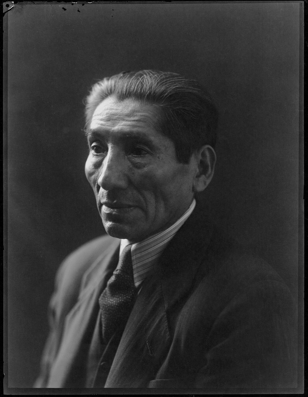 Black-and-white self-portrait of Martín Chambi in a dark suit, striped shirt, and tie, looking slightly to one side.