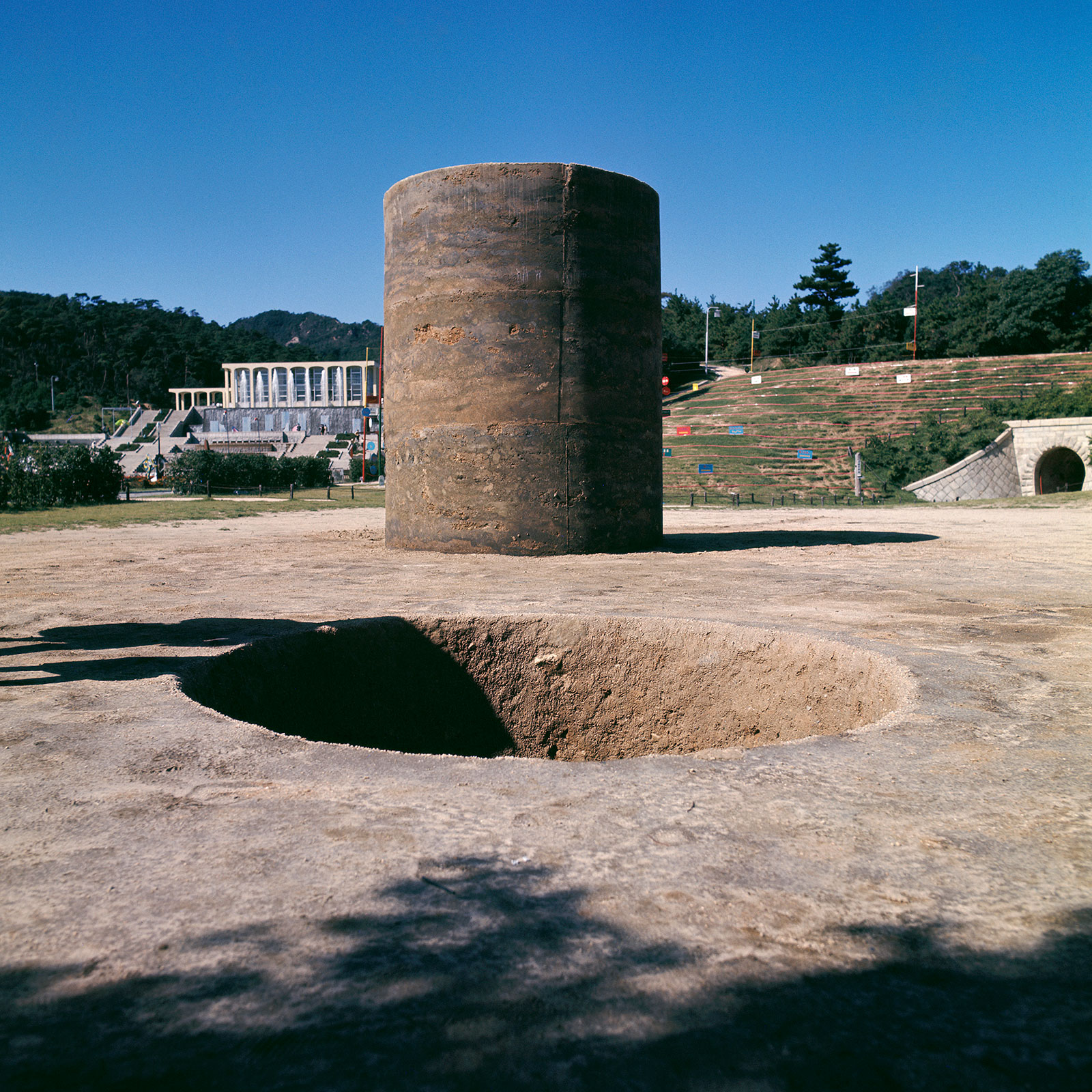 Archival installation photograph of Nobuo Sekine's Phase—Mother Earth, showing a deep circular excavation beside a matching upright cylinder of compacted earth at Kobe Suma Rikyū Park.