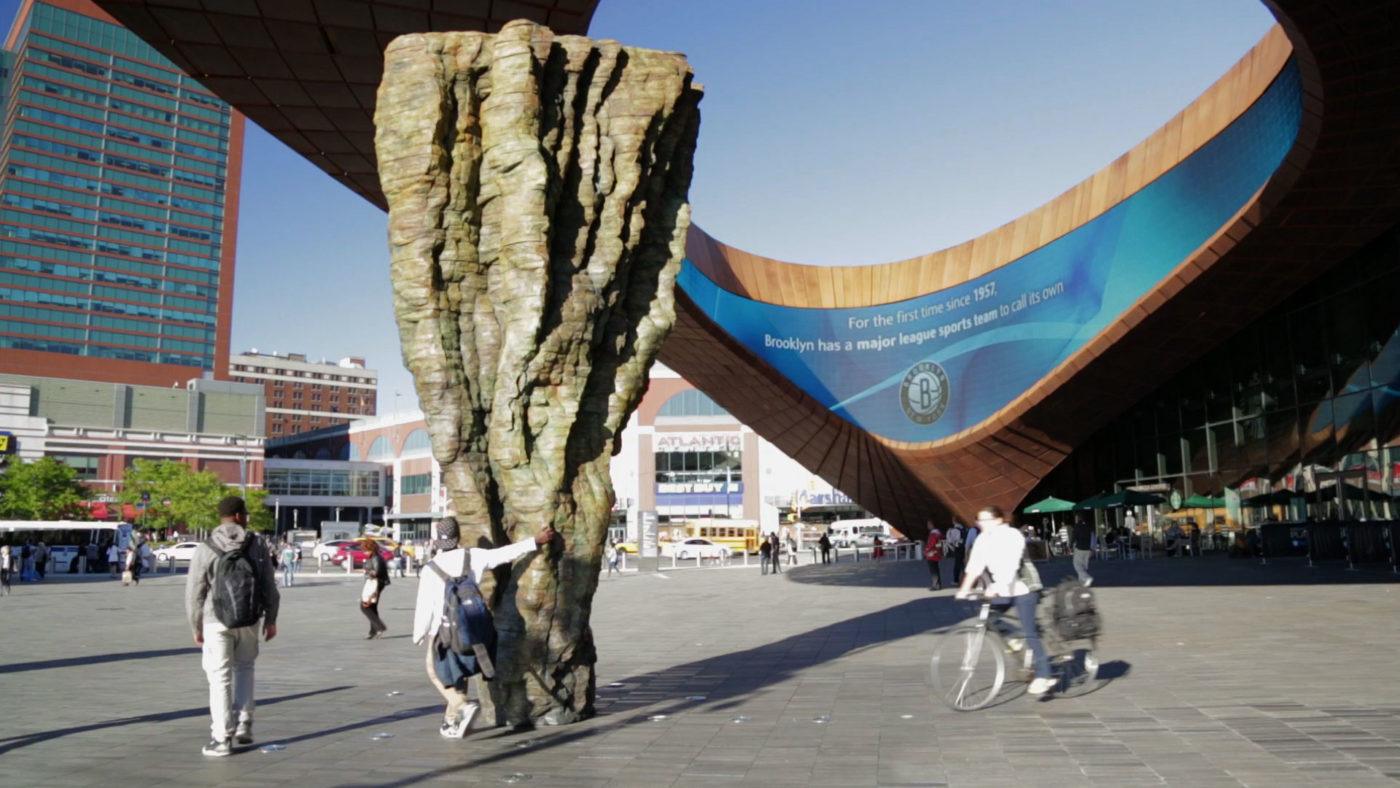 Ursula von Rydingsvard's tall bronze sculpture Ona stands in the Barclays Center plaza as pedestrians walk past and one visitor touches its deeply ridged surface.