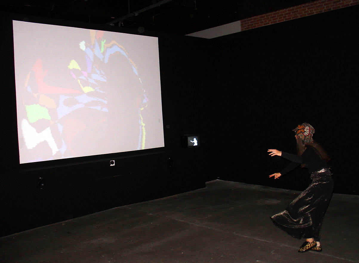 Galen Brandt bends and extends both arms toward a large VIDEOPLACE projection in a dark gallery, where layered colored silhouettes appear on the screen.