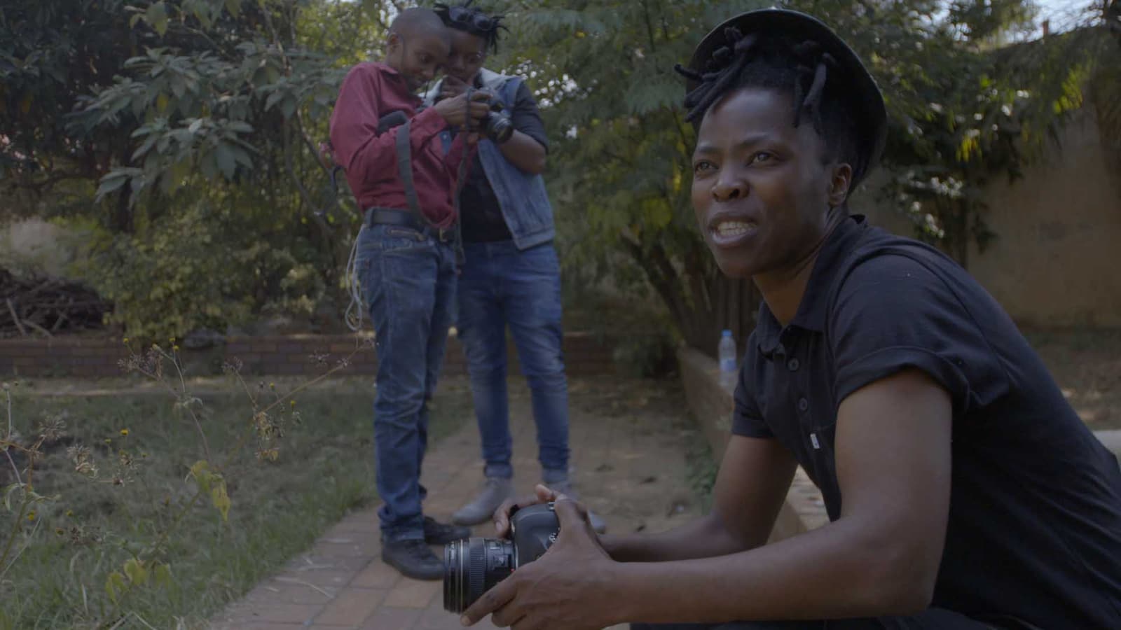 Zanele Muholi crouches outdoors holding a DSLR while two collaborators review another camera in Daveyton, South Africa.