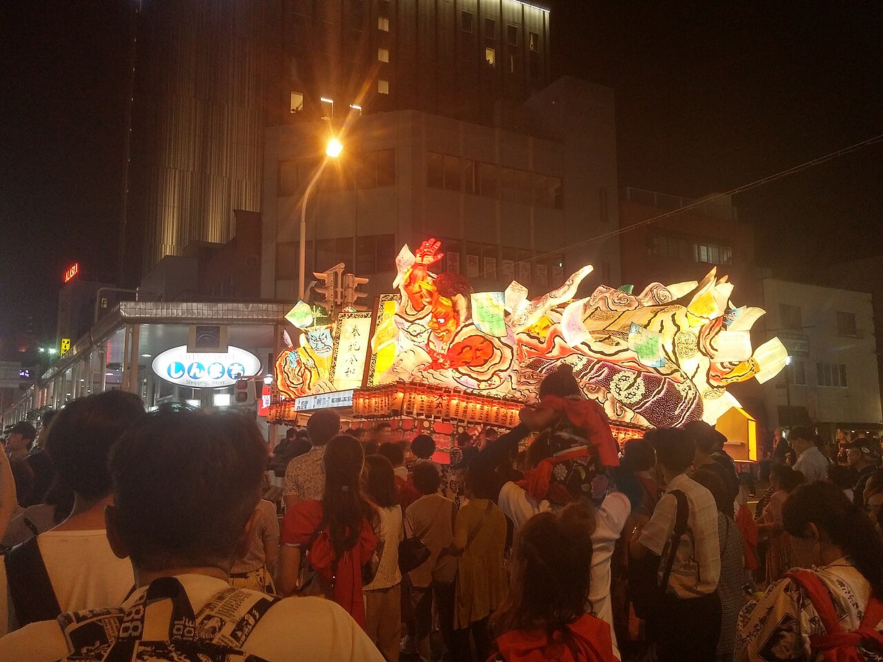 一辆点亮的青森睡魔灯车穿过新町通夜间密集的人群。