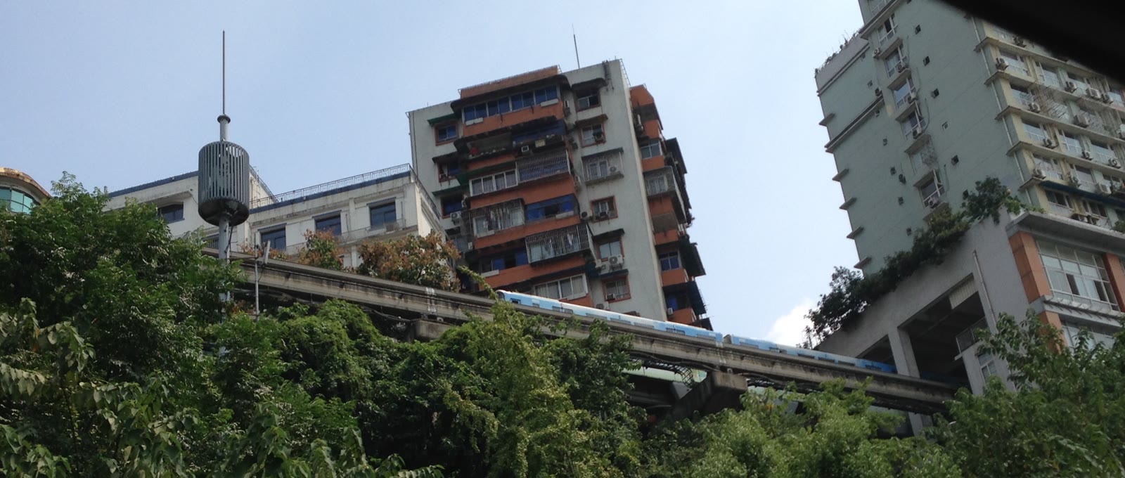 A blue Line 2 monorail train passing through the building that contains Liziba Station above a steep green slope in Chongqing.