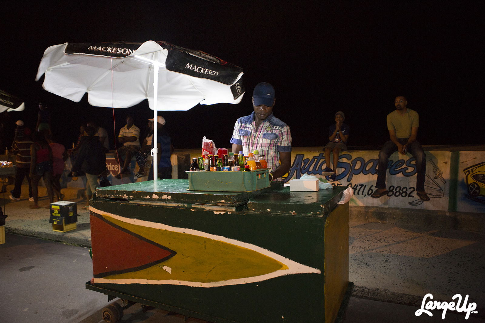 A vendor behind a wheeled cart painted green, yellow, red, and black while people sit on Georgetown’s Sea Wall at night.