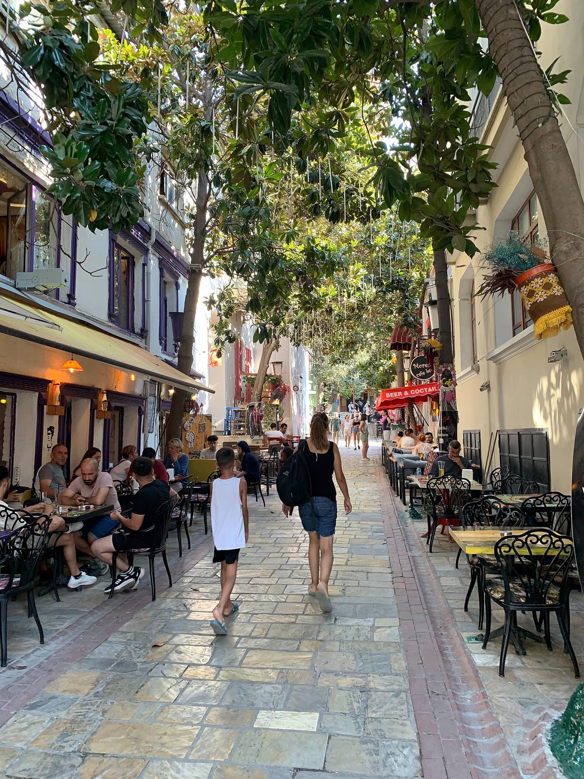 People walking and sitting at cafe tables beneath trees on the narrow stone-paved Dario Moreno Street in İzmir.