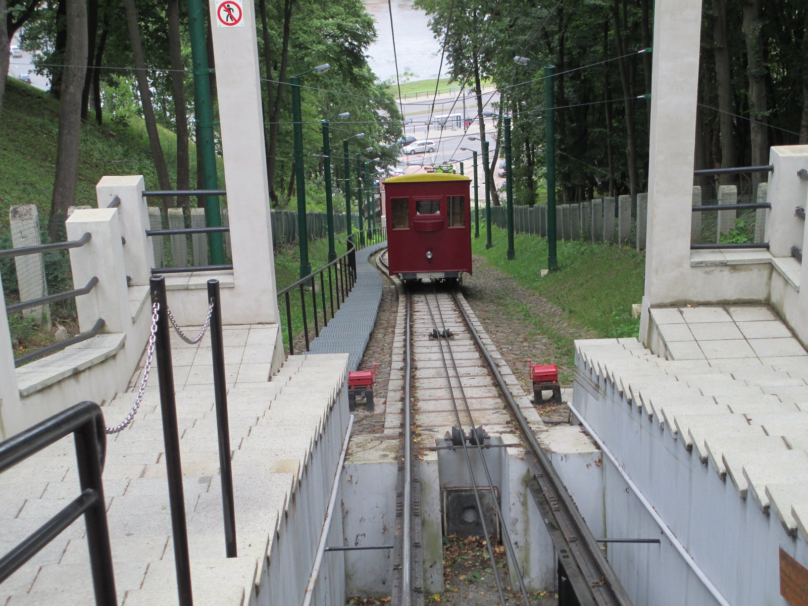 A red Aleksotas Funicular car descending a steep, tree-lined track toward the Nemunas in Kaunas.