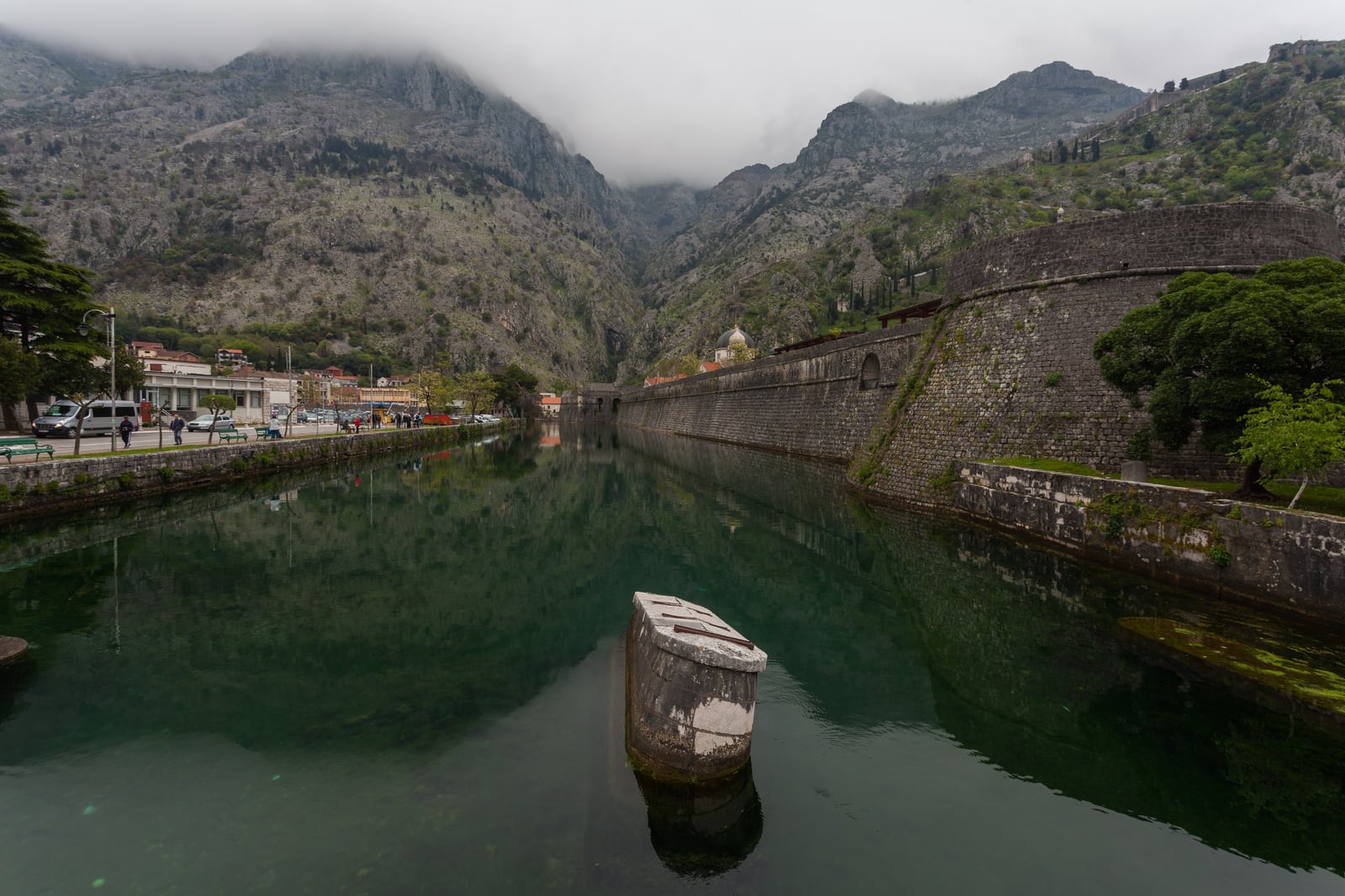 The reflective green Škurda River beside Kotor's long stone wall and rounded Kampana bastion, backed by cloud-covered mountains.