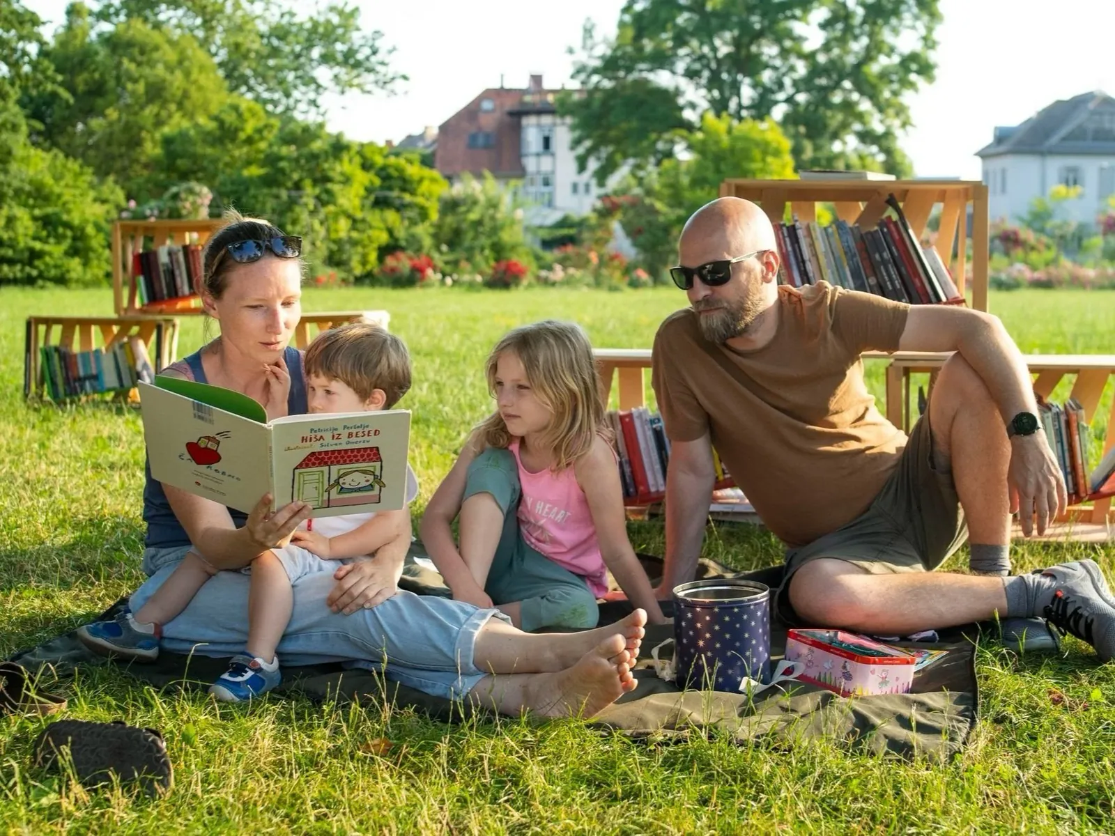 Two adults and two children sitting on a blanket with an open picture book on a sunlit lawn, with wooden book crates from Ljubljana’s Library Under the Treetops behind them.