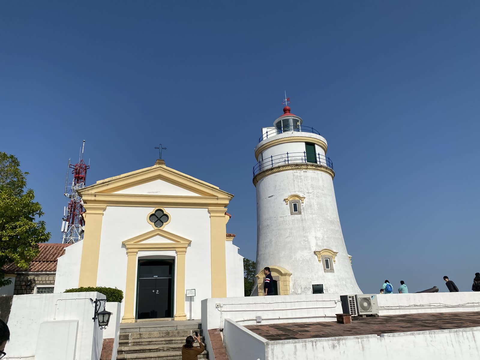 Guia Chapel and Guia Lighthouse on Guia Hill in Macao, with the white chapel wall, lighthouse tower, trees, and paved terrace visible.