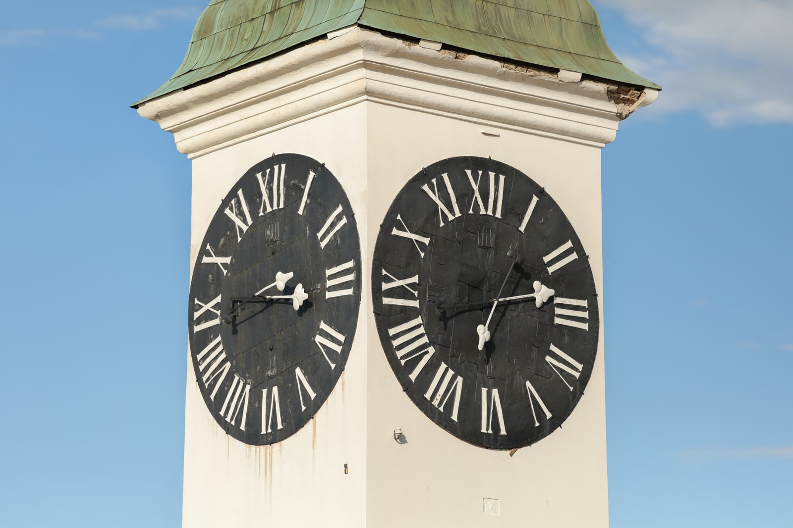 Two black Roman-numeral faces of Petrovaradin's white clock tower, with long hands indicating the hour and short hands indicating the minutes beneath a weathered green roof.