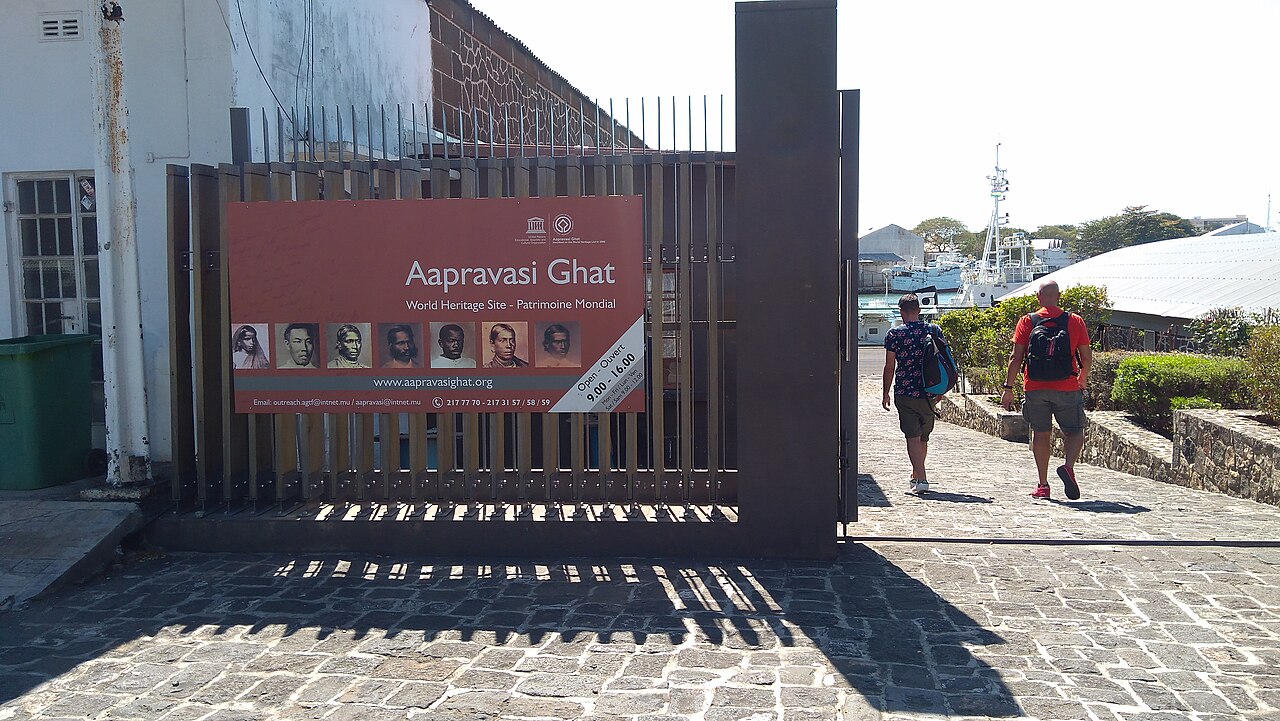 Two visitors walk toward the Aapravasi Ghat museum entrance in Port Louis, with a heritage sign, cobbled ground, and harbor vessels behind them.