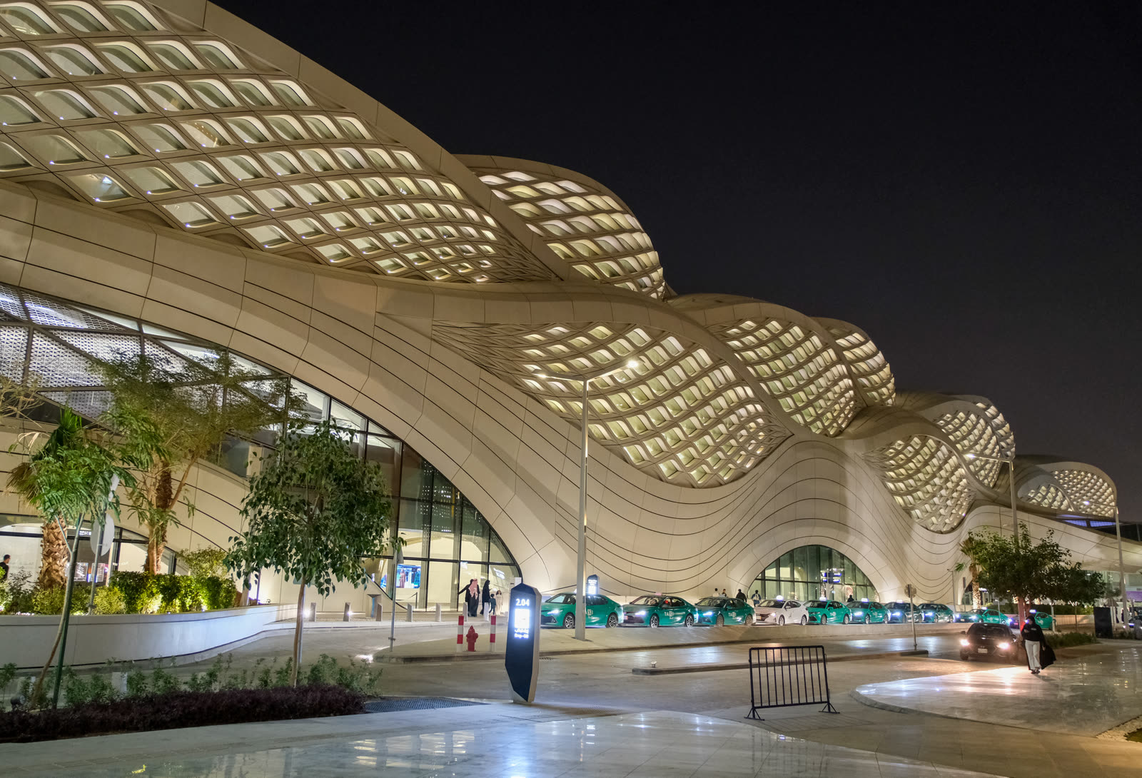 KAFD Metro Station in Riyadh at night, with its illuminated undulating facade, taxi lane, station entrance, and palm planting visible.