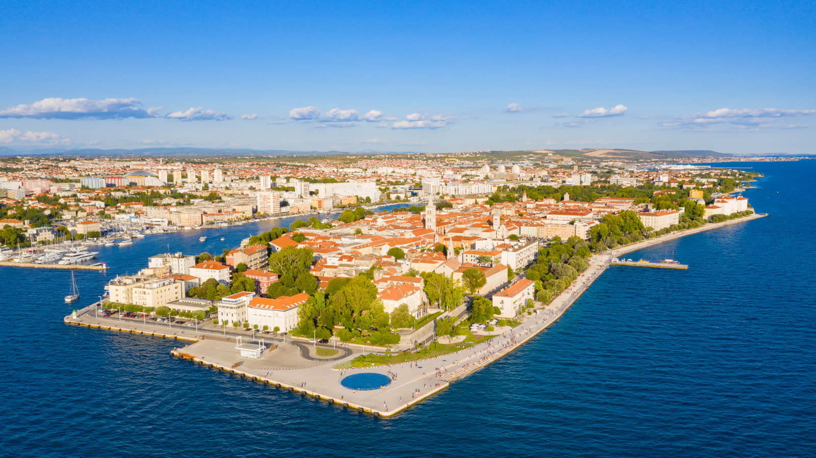 Aerial view of Zadar's waterfront showing the circular Greeting to the Sun installation beside the stepped Sea Organ and the Adriatic water.
