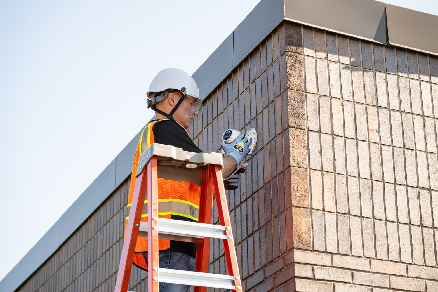 A safety technician in a hard hat and reflective vest inspects a wall-mounted fire-safety device from a ladder.