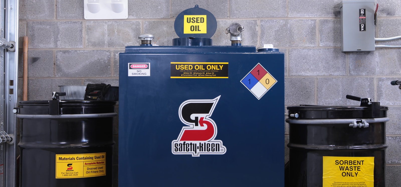 A dark-blue Safety-Kleen used-oil collection tank between two black waste drums in a service bay.