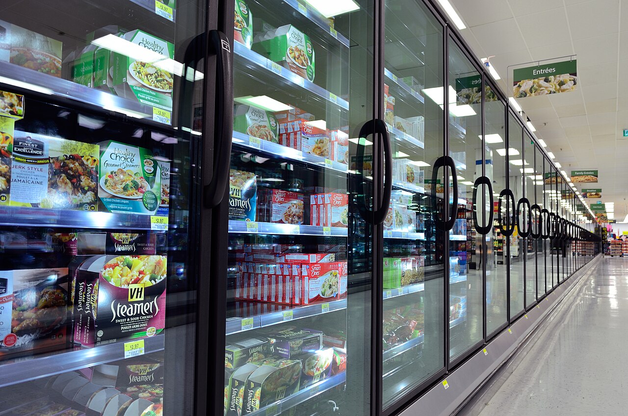 A supermarket frozen-food aisle with glass freezer doors, boxed meals, and frozen products visible under store lighting.